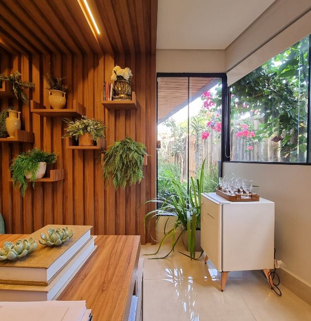 Sala decorada com parede de madeira, prateleiras com plantas, mesa de madeira e vista para um jardim com flores e vegetação